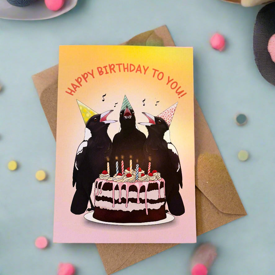 Birthday card with three magpies at a cake, surrounded by flowers and an envelope.